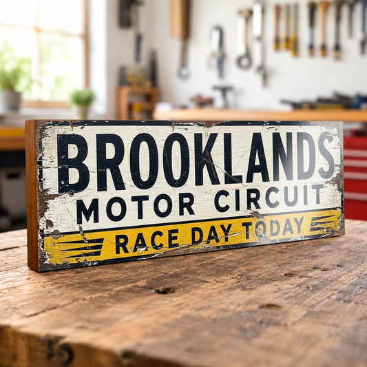 A rectangular wooden sign with a distressed vintage design reading 'BROOKLANDS MOTOR CIRCUIT' and 'RACE DAY TODAY,' displayed on a rustic wooden table in a blurred garage or workshop setting.