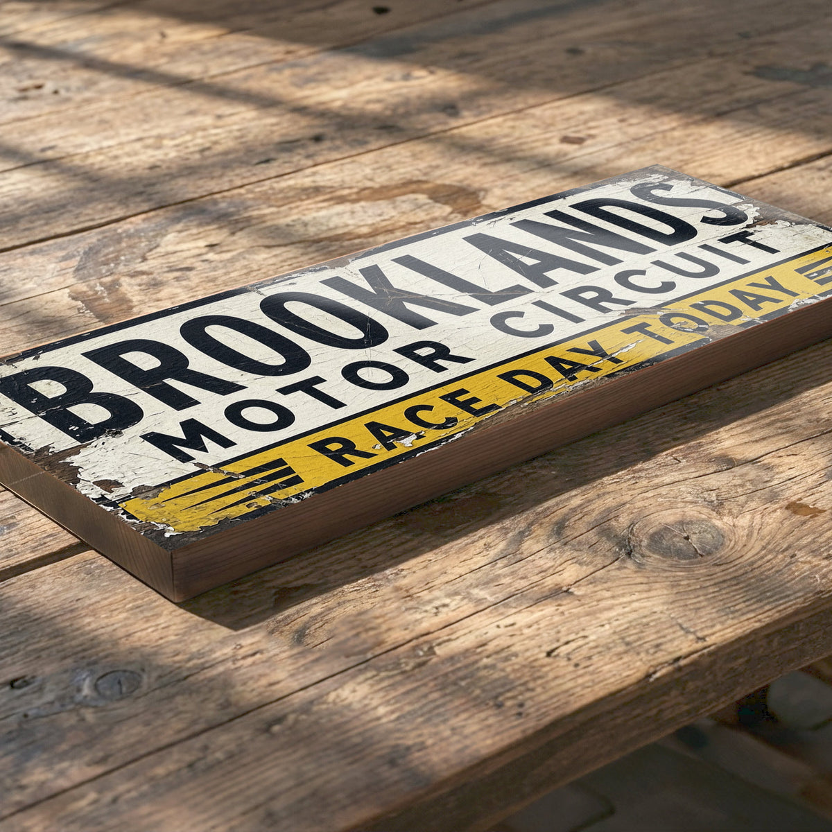 A rectangular wooden sign with a distressed vintage design reading 'BROOKLANDS MOTOR CIRCUIT' and 'RACE DAY TODAY', placed on a rustic wooden table.