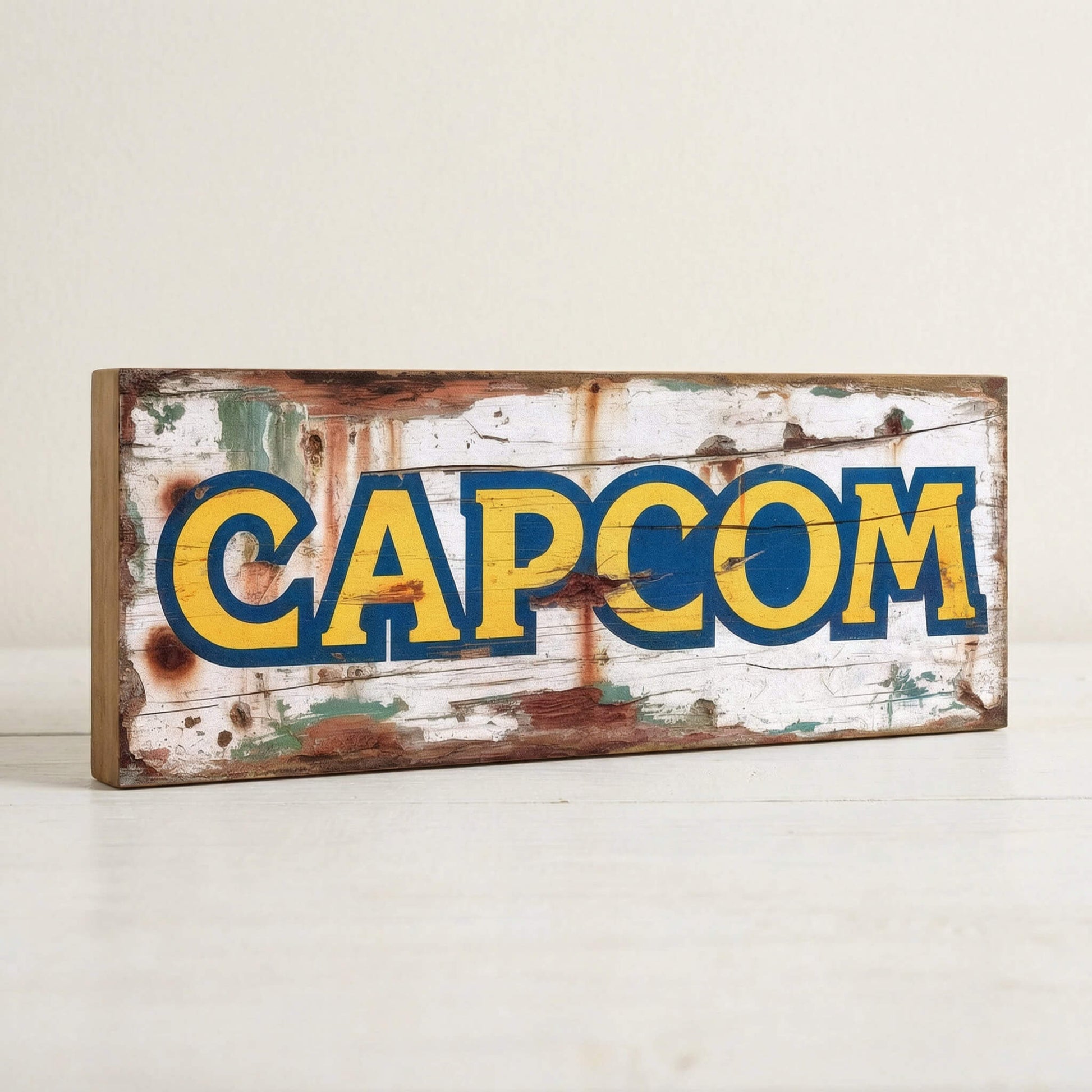 Capcom retro gaming logo vintage wooden sign handmade on 18mm MDF with bold yellow and blue lettering on heavily distressed white background, freestanding on light surface