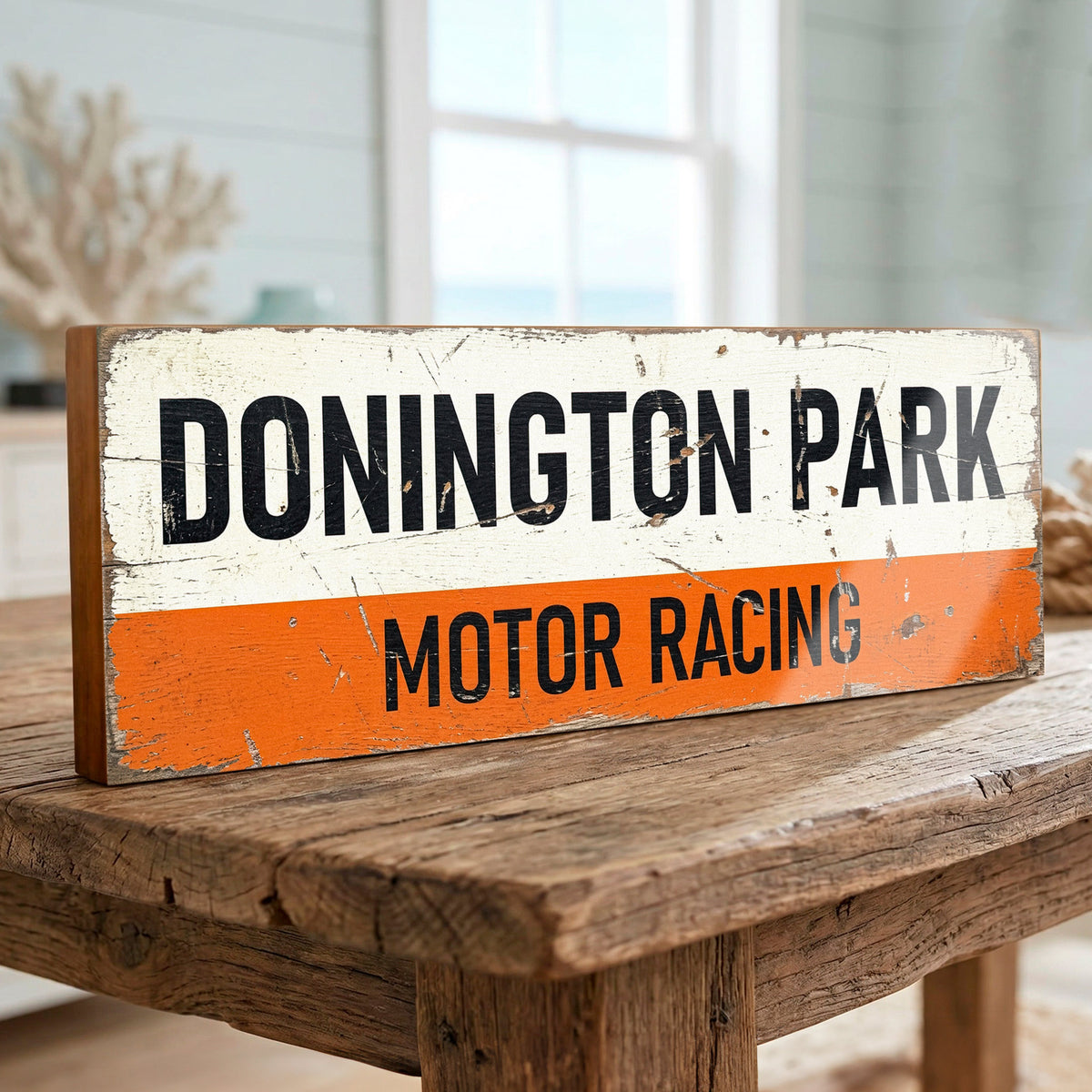 A rustic rectangular wooden sign reading "DONINGTON PARK MOTOR RACING" in black lettering, with a distressed white top half and orange bottom half, displayed on a wooden table in a softly blurred indoor setting.