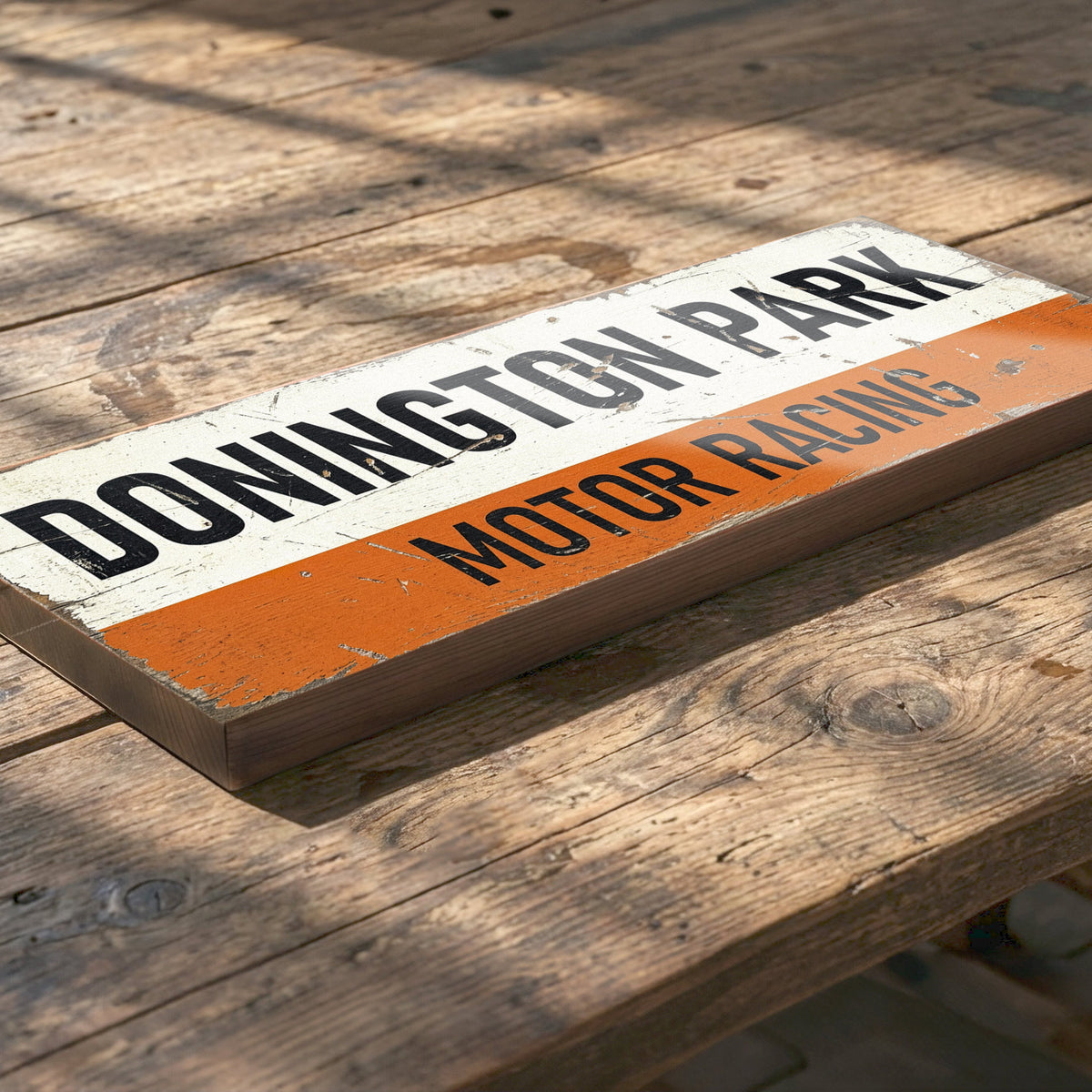 A rectangular wooden sign with a distressed finish, featuring the text 'DONINGTON PARK MOTOR RACING' in black lettering across an off-white top section and an orange bottom stripe. The sign is photographed at an angle on a rustic wooden plank surface.