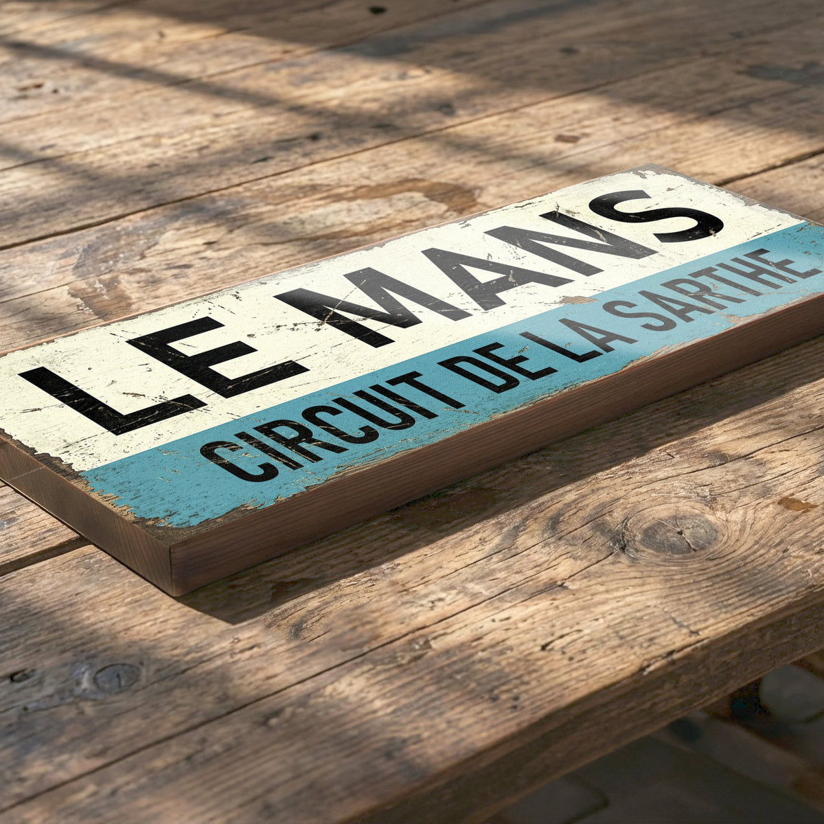 A rectangular wooden sign with a distressed finish, featuring the text 'LE MANS' in black on an off-white top section and 'CIRCUIT DE LA SARTHE' in black on a light blue bottom section. The sign is placed diagonally on a rustic wooden plank surface.