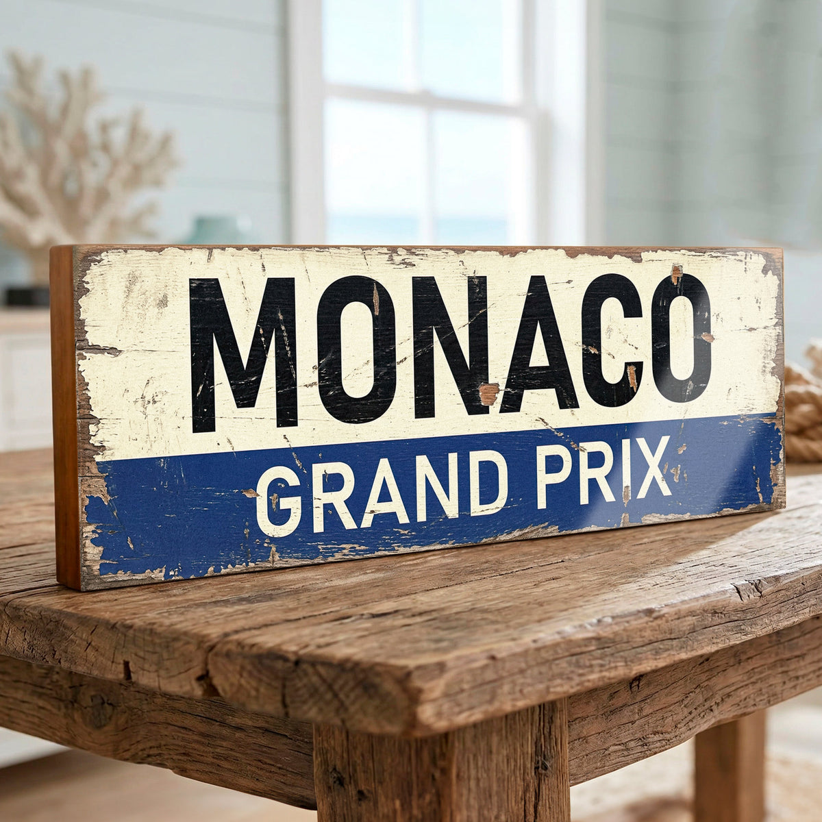 A rectangular distressed sign reading 'MONACO GRAND PRIX' in black and white lettering on an off-white top section and blue bottom stripe, displayed on a rustic wooden table in a softly blurred indoor setting.