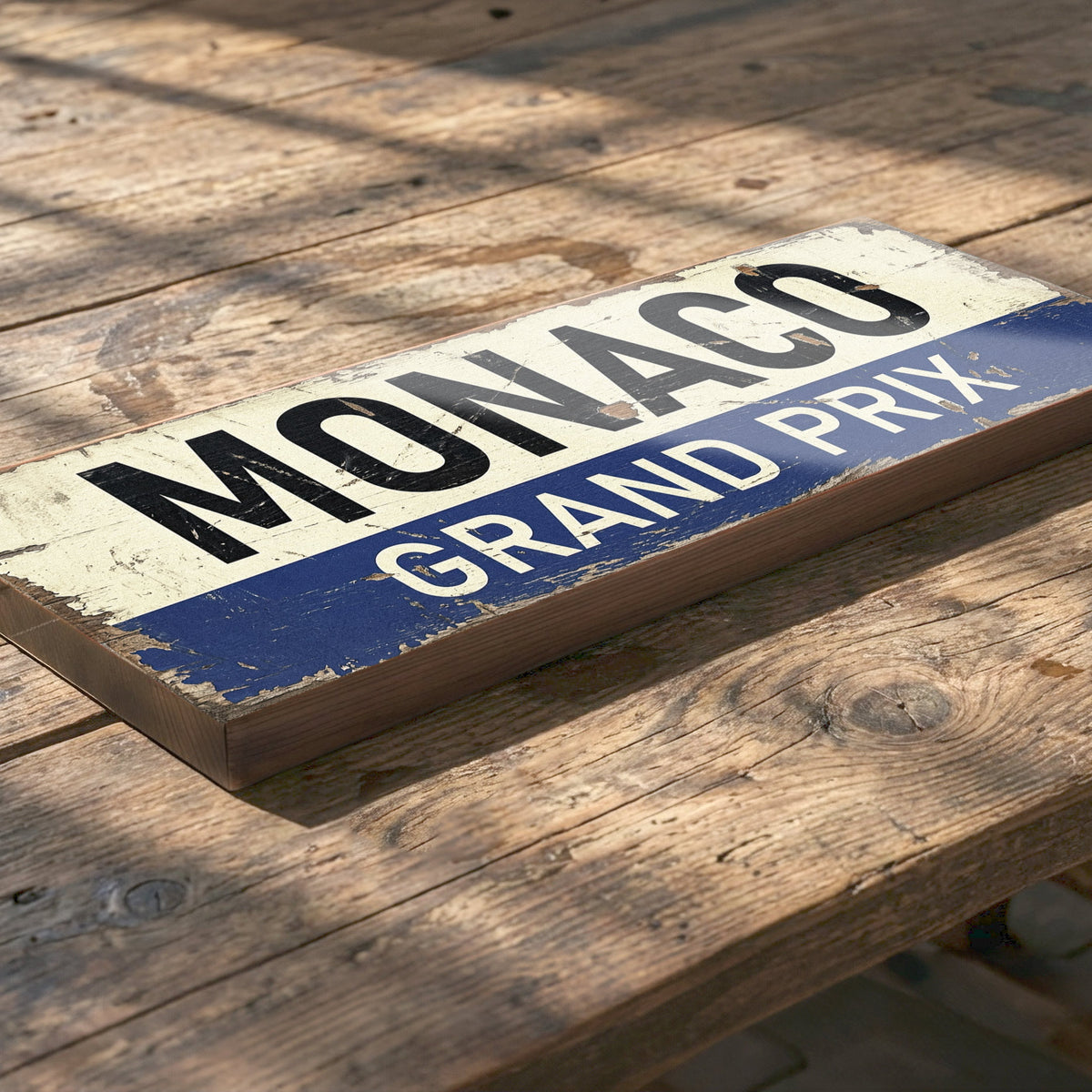 A rectangular wooden sign reading 'MONACO GRAND PRIX' in distressed blue and cream colors, placed on a rustic wooden table.