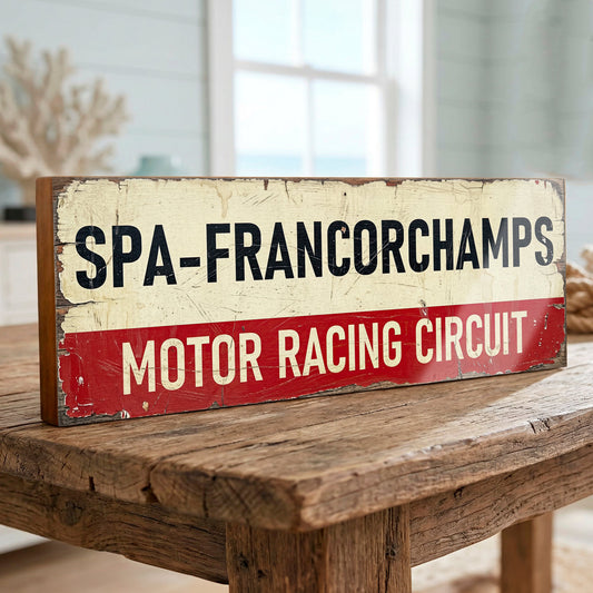 A rustic rectangular wooden sign reading 'SPA-FRANCORCHAMPS MOTOR RACING CIRCUIT' in black and cream lettering on a red lower band, displayed on a wooden table in a softly blurred indoor setting.