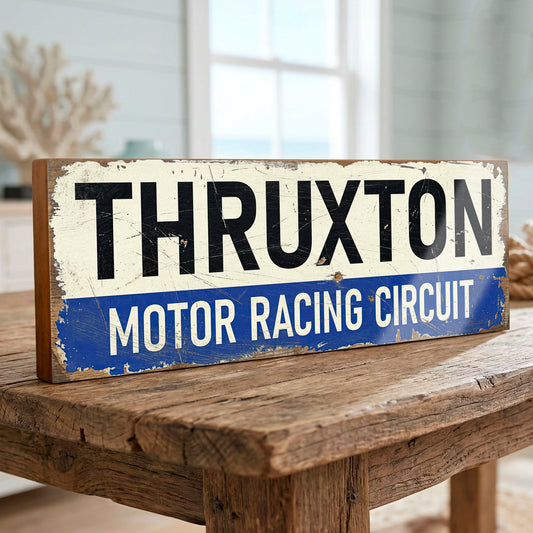 A rustic rectangular metal sign reading 'THRUXTON MOTOR RACING CIRCUIT' in black and white lettering on a distressed cream and blue background, displayed on a wooden table in a softly blurred indoor setting.