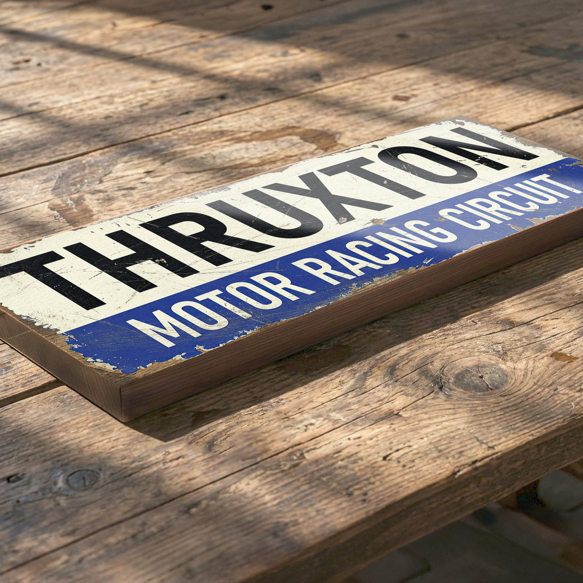 A rectangular wooden sign with a distressed vintage design featuring the text 'THRUXTON MOTOR RACING CIRCUIT' in black and white on a blue band, placed on a rustic wooden table.