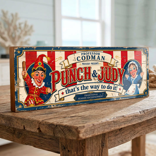 A rectangular vintage-style wooden sign featuring the text 'Professor Codman proudly presents Punch & Judy that's the way to do it!' with illustrated clowns, displayed on a rustic wooden table in a softly blurred indoor setting.