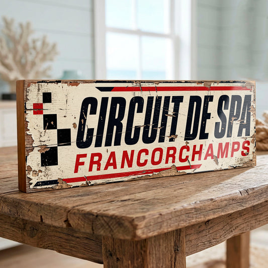 A rustic rectangular wooden sign reading 'Circuit de Spa Francorchamps' in bold black and red lettering on a distressed cream background, displayed on a wooden table in a softly blurred indoor setting.